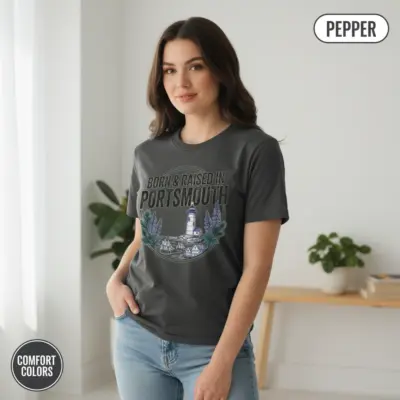 Young woman wearing a pepper Comfort Colors t-shirt with Born and Raised in Portsmouth 603 NH city pride graphic in casual indoor setting