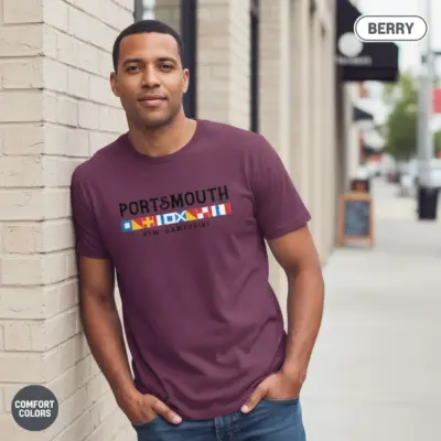 Man wearing a berry Comfort Colors t-shirt featuring Portsmouth New Hampshire spelled in international nautical signal flags on a city street