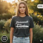 Woman wearing a pepper-colored garment-dyed t-shirt featuring Portsmouth NH coordinates 43.07°N 70.76°W with a minimalist waterfront skyline illustration, photographed outdoors in natural light