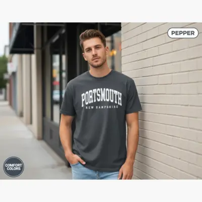 Young man wearing a pepper black Comfort Colors t-shirt featuring a Portsmouth NH varsity-style hometown pride graphic on the front chest