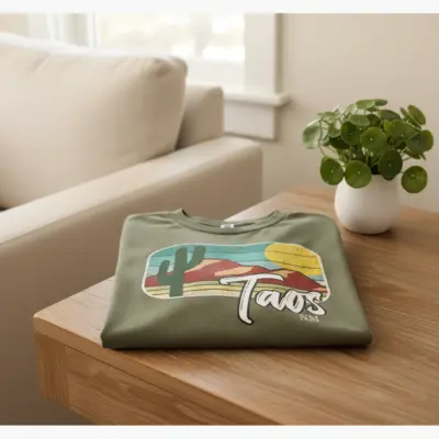 Moss green Comfort Colors t-shirt featuring a Taos New Mexico high desert sagebrush and Sangre de Cristo mountain landscape graphic laid flat
