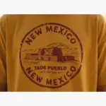 Mustard garment-dyed Comfort Colors t-shirt closeup showing Taos New Mexico souvenir graphic with mountain and adobe design detail