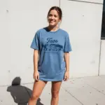 Woman wearing a Blue Jean Comfort Colors garment-dyed t-shirt featuring the Taos NM coordinates 36.41°N 105.57°W in a minimalist front-chest print, photographed in natural sunlight