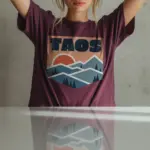 taos-skyline-shirt-adobe-mountain-new-mexico-tee-comfort-colors-t-shirt-creative-female-berry.webp Woman wearing a berry Comfort Colors t-shirt featuring a minimalist Taos New Mexico skyline illustration with adobe architecture and Wheeler Peak