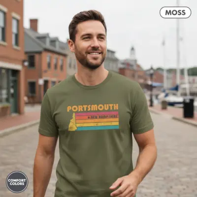 Man wearing moss green vintage-style Portsmouth New Hampshire seaport graphic t-shirt in an outdoor coastal setting